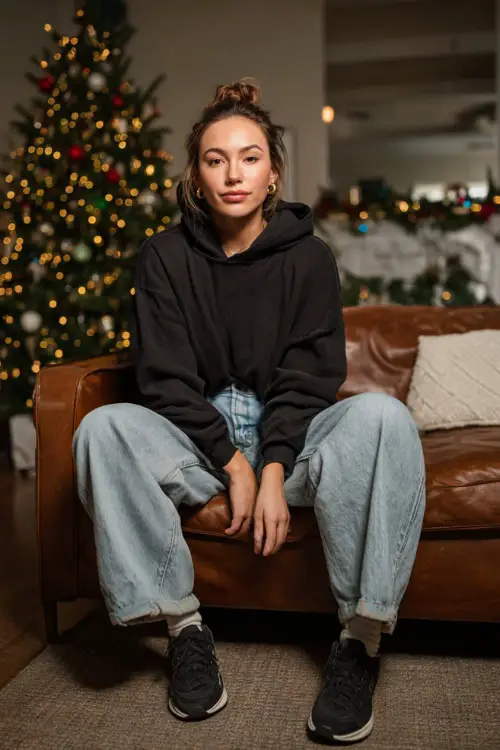 A woman wears a black hoodie, baggy light-wash denim, black sneakers, simple cozy Christmas living room with couch and tree, full body portrait