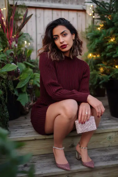A woman wears a chic burgundy ribbed sweater dress, small crystal clutch, strappy block heels, winter photoshoot set with Christmas plants and soft warm lights