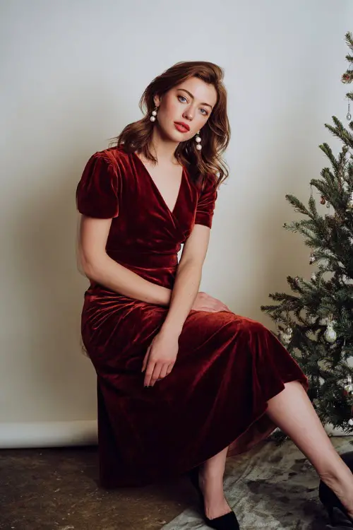 A woman wears a classic red velvet midi dress, pearl earrings, black heels, white photography studio with a small decorated Christmas tree prop and soft light shadows
