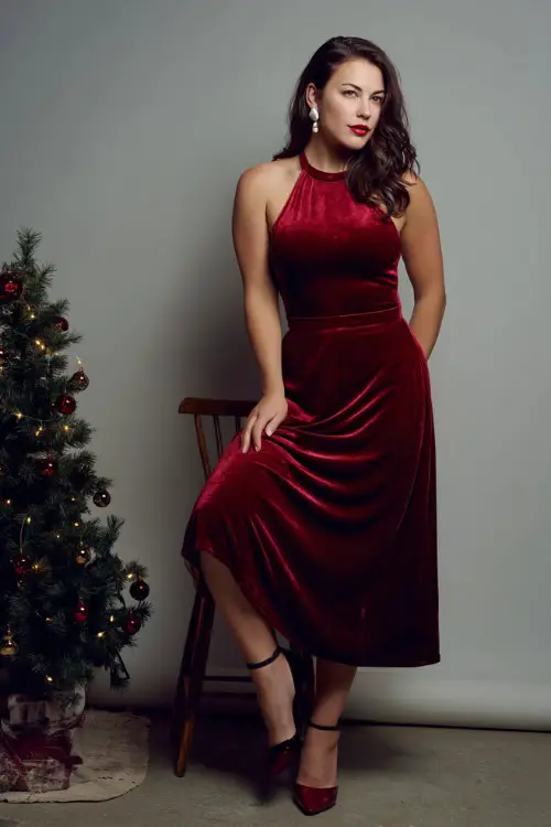 A woman wears a classic red velvet midi dress, pearl earrings, black heels, white photography studio with a small decorated Christmas tree prop and soft light shadows 