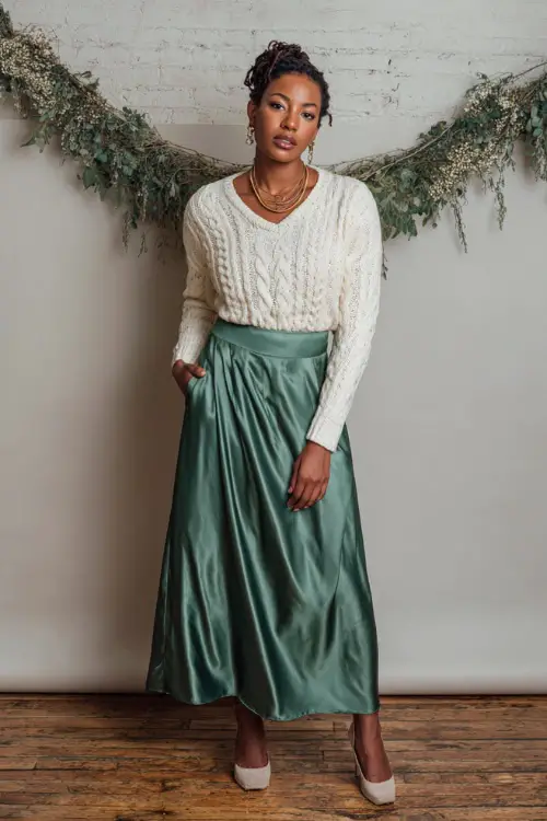 A woman wears a cream cable knit sweater tucked into an elegant satin green maxi skirt, gold delicate necklace, ivory heels