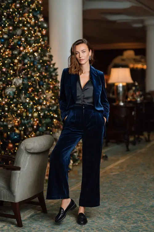 A woman wears a navy velvet blazer set with wide-leg trousers, silk blouse, and black loafers, chic winter party environment with a grand Christmas tree behind 