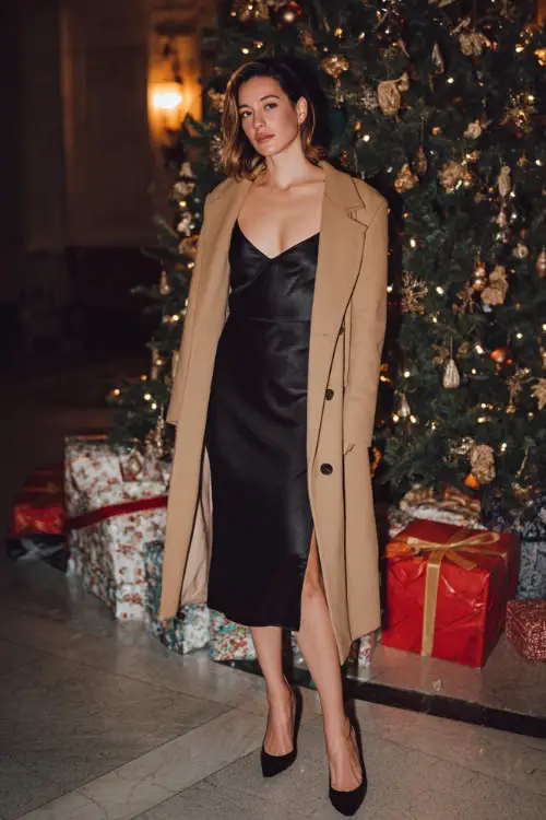 A woman wears a timeless black satin slip dress layered with a long camel coat, pointed heels, dim elegant lighting photoshoot in front of a grand Christmas tree with wrapped gift boxes 