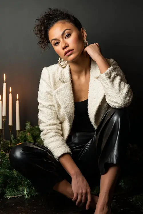 A woman wears an ivory boucle jacket, black high-rise pants, pearl earrings and leather heels, candle-lit photoshoot setup with Christmas greenery props