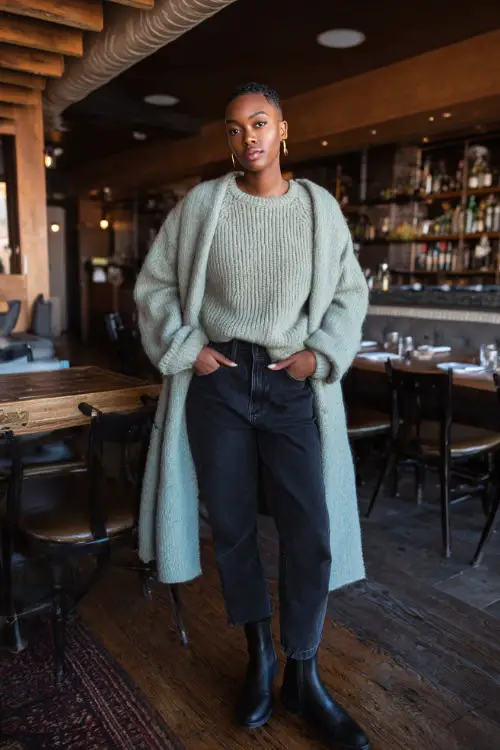 A Black woman dressed in straight-leg jeans with a soft knit sweater and a long wool coat, styled with Chelsea boots and minimal accessories, posing in a calm winter brunch environment