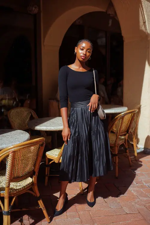 A Black woman in a soft knit top paired with a pleated midi skirt and ballet flats, styled with a crossbody bag, posing near café tables under morning sunlight