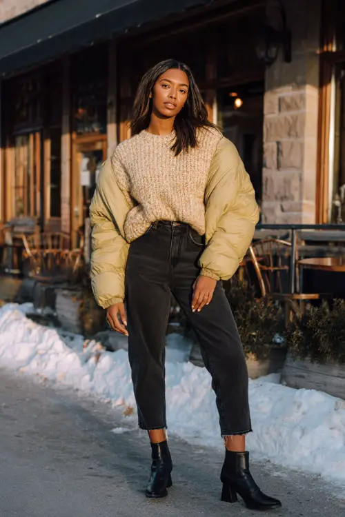 A Black woman wears a cozy oversized knit sweater with dark jeans, heeled booties, and a stylish puffer jacket, posing near a winter café with warm glowing windows
