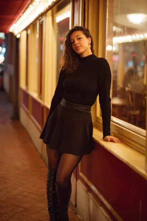 A curvy woman wears a ribbed knit sweater with a black A-line midi skirt, sheer tights, and knee-high leather boots