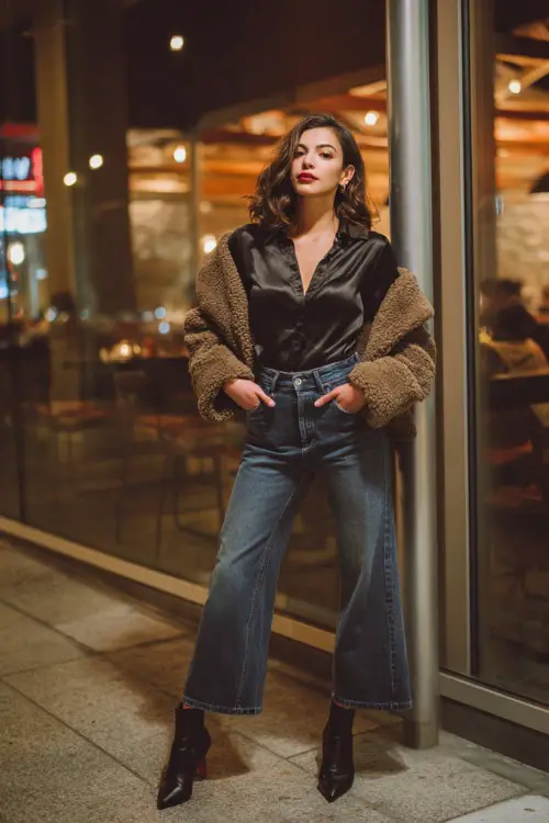 A midsize woman wears a black satin blouse with wide-leg jeans, styled with heeled booties and a cozy shearling jacket