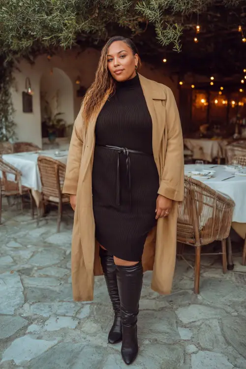 A plus-size woman over 30 wears a black knit midi dress with a belted waist, paired with knee-high leather boots and a camel coat, posing beside a softly lit winter dinner venue
