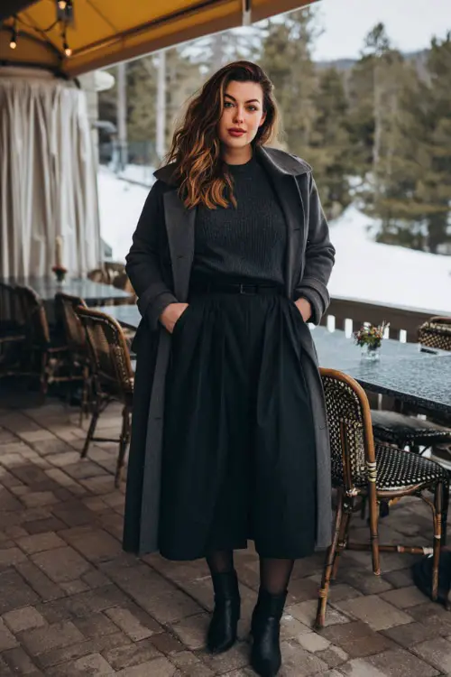 A plus-size woman wears a chic charcoal sweater tucked into a pleated black midi skirt, paired with tights, ankle boots, and a belted wool trench