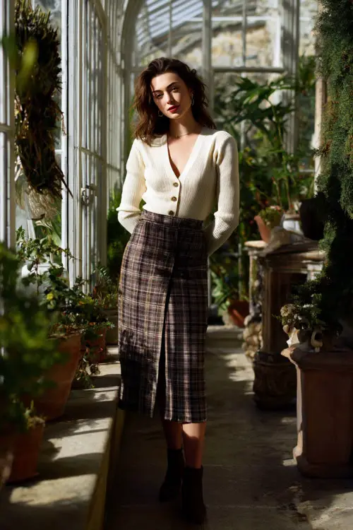 A refined woman over 30 wears a plaid midi skirt with a cream cardigan and heeled boots, standing full body in a sunlit conservatory filled with winter greenery