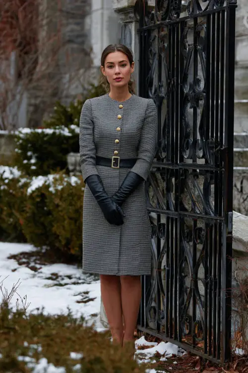 A refined woman wears a gray tailored dress with gold buttons and a matching structured coat, paired with black leather gloves and heels