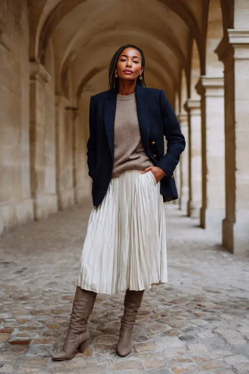 A stylish woman over 30 wears a cream pleated skirt with a taupe cashmere sweater and a tailored navy jacket, styled with heeled boots