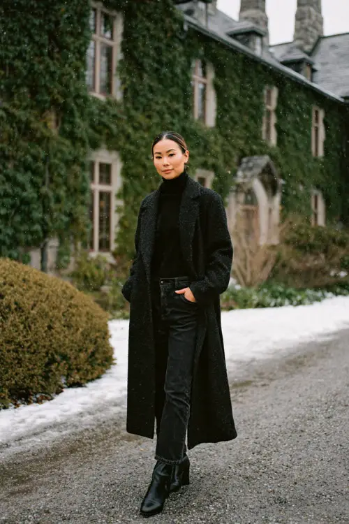 A stylish woman over 30 wears tailored black jeans with a charcoal turtleneck, a long black wool coat, and polished ankle boots