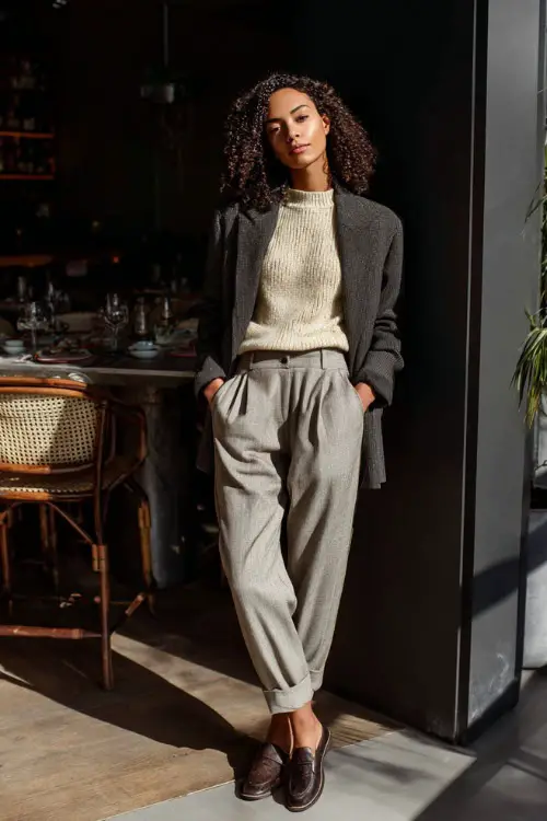 A woman dressed in a knit sweater layered under a tailored blazer and wool trousers, paired with loafers or low heels, standing in a chic indoor brunch café with natural light