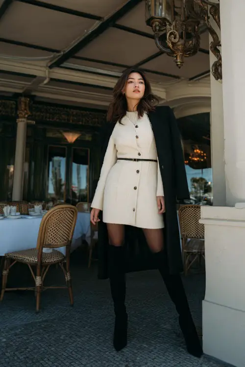 A woman dressed in a structured mini-length winter dress styled with tights, knee-high boots, and a tailored coat, posing outside a fashionable winter brunch location