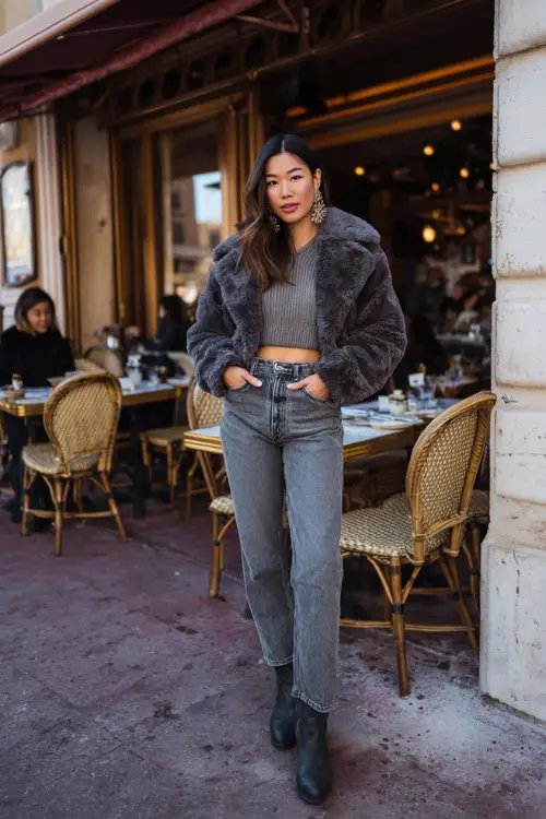 A woman dressed in straight-leg jeans with a chic knit top and a cropped wool jacket, styled with ankle boots and statement earrings, posing outside a busy winter brunch café