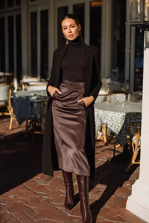 A woman in a fitted turtleneck tucked into a satin midi skirt, layered with a long elegant coat and heeled ankle boots