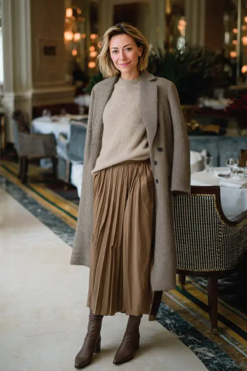 A woman in a soft cashmere sweater tucked into a pleated midi skirt, layered with a structured coat and ankle boots, posing in an elegant birthday brunch environment during winter