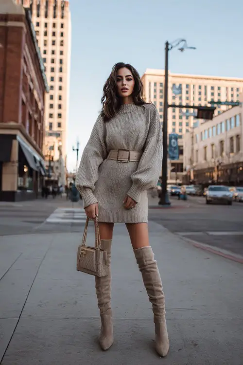 A woman in a sweater dress cinched at the waist with a belt, paired with knee-high boots and a structured handbag, posing on a city street during winter brunch hours