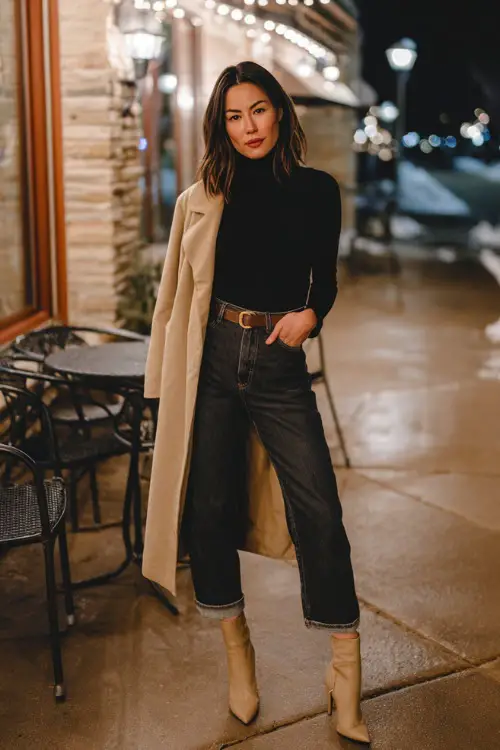 A woman over 30 wears a fitted black mock-neck top with straight-leg jeans, heeled booties, and a belted camel coat, posing beside a softly lit winter café during date night 