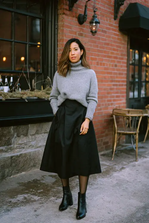 A woman over 30 wears a soft gray sweater tucked into a black A-line midi skirt, styled with tights and ankle boots, standing outside a relaxed winter dinner venue