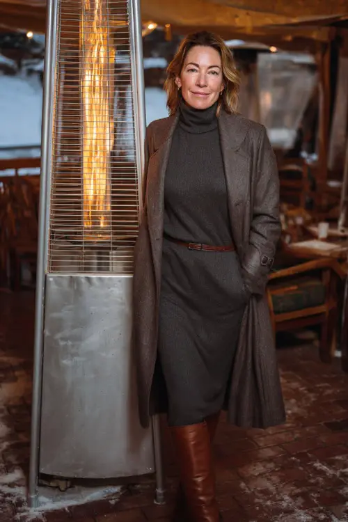 A woman over 40 wears a charcoal turtleneck dress under a belted wool trench coat, styled with leather knee boots, standing near a winter patio heater outside a dinner spot 