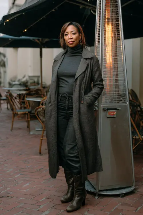 A woman over 40 wears a charcoal turtleneck tucked into black jeans, styled with leather boots and a chic wool trench coat