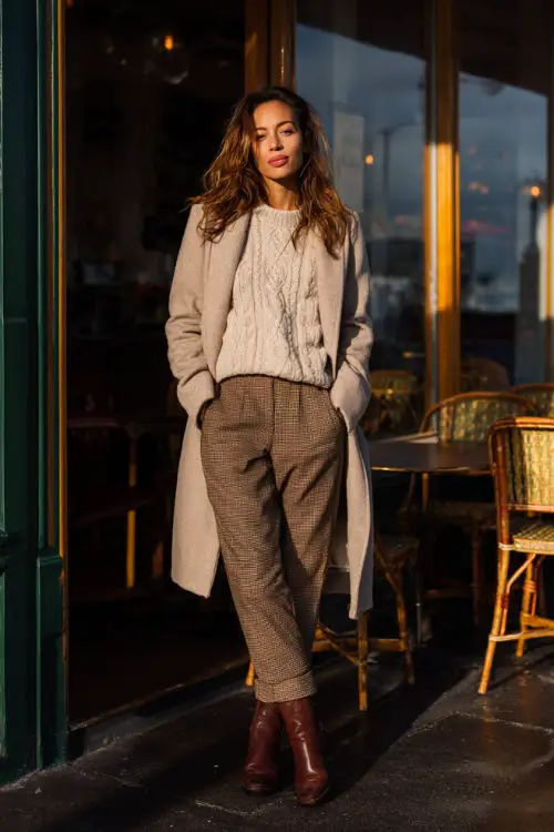 A woman wearing a cozy knit sweater tucked into tailored trousers, layered with a long wool coat and styled with ankle boots, standing outside a winter brunch café in soft daylight