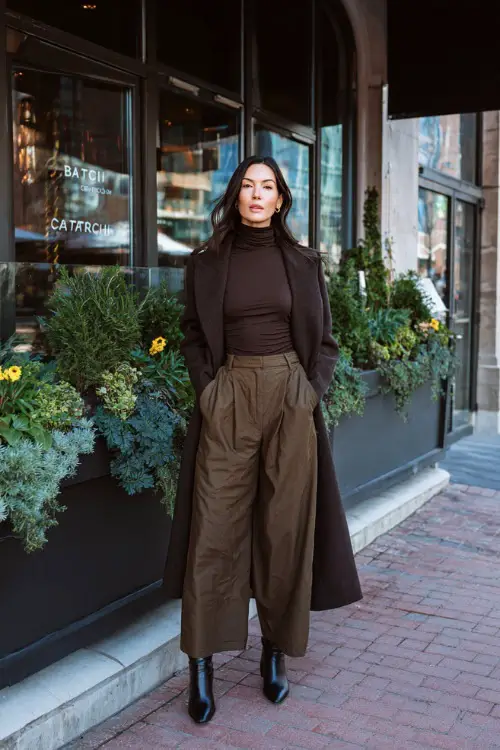A woman wearing a fitted turtleneck tucked into wide-leg trousers, layered with a long elegant coat and heeled ankle boots