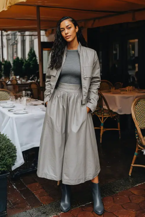 A woman wearing a long-sleeve midi skirt paired with a fitted knit top and a cropped waterproof jacket, styled with ankle boots