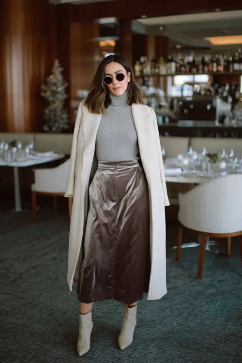A woman wearing a sleek knit top paired with a satin or velvet midi skirt, layered with a long coat and elegant ankle boots, standing in a chic winter birthday brunch setting 