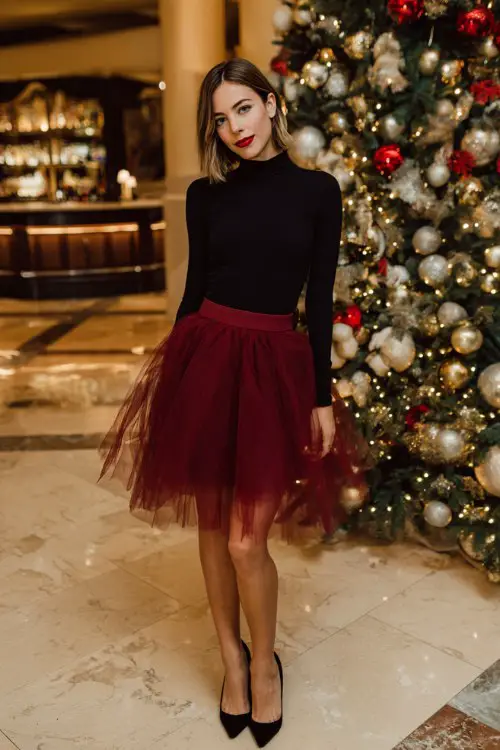 A woman wears a black long-sleeve fitted top with a ruby tulle skirt, sleek heels, full body at a festive holiday event with ornaments and a glowing Christmas tree
