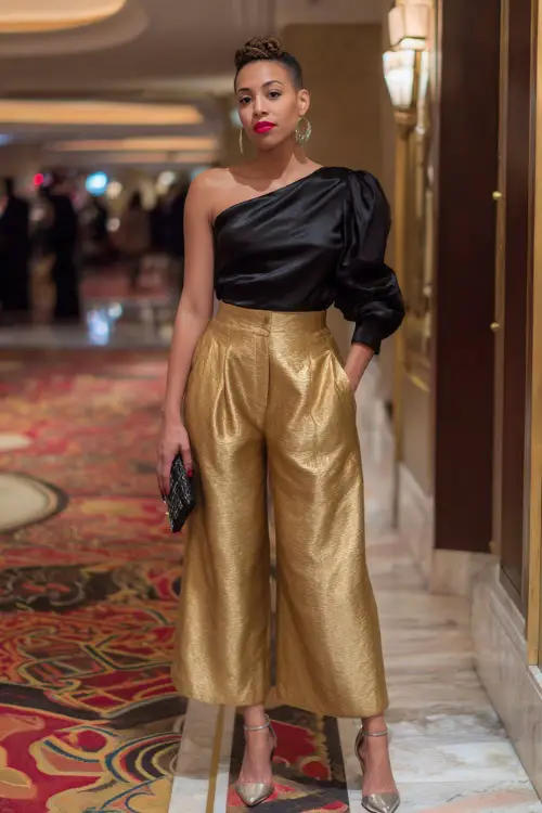 A woman wears a black satin one-shoulder blouse, gold wide-leg pants, metallic heels, full outfit visible in a glamorous hotel holiday party hall