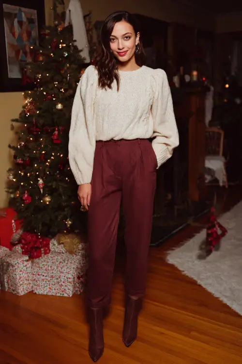 A woman wears a cream knit sweater tucked into burgundy straight-leg pants, ankle boots with a heel, full body framing at a cozy house Christmas party with warm décor