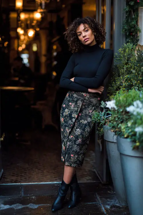 A woman wears a fitted long-sleeve top tucked into a dark floral midi skirt, paired with heeled boots and a tailored coat, posing near a softly lit winter restaurant entrance