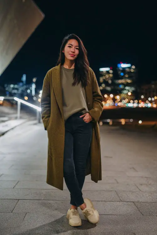 A woman wears a forest-green sweatshirt with a long wool coat, slim dark jeans, and platform sneakers, posing near a modern winter city plaza at night