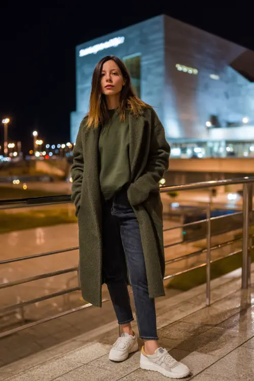 A woman wears a forest-green sweatshirt with a long wool coat, slim dark jeans, and platform sneakers, posing near a modern winter city plaza at night