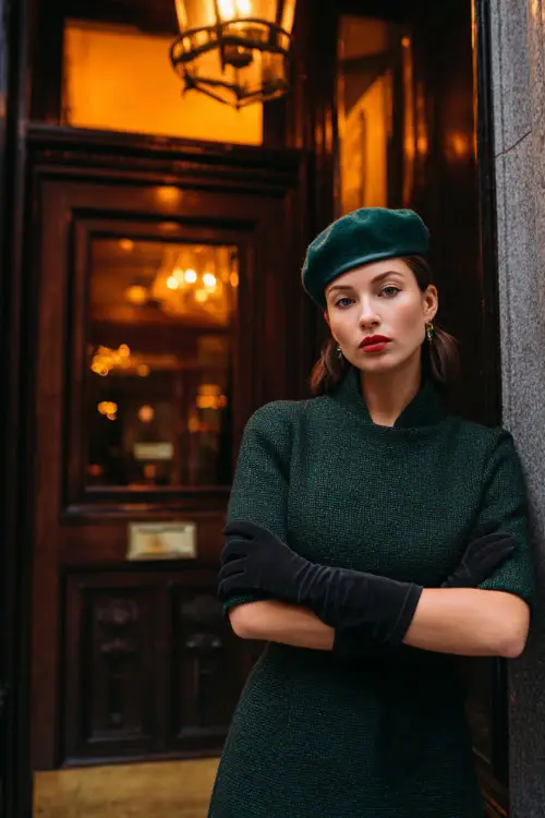 A woman wears a forest green tweed dress with a fitted waist, matching beret, and long black gloves, full body in front of a private members club with warm light glowing from inside 