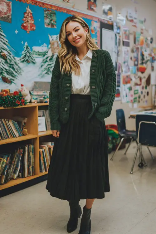 A woman wears a green cable-knit cardigan over a white blouse, pleated black midi skirt, and comfortable block-heel boots
