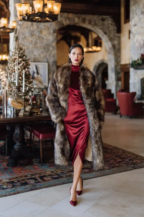 A woman wears a high-neck long-sleeve satin dress in deep red, layered with a fur-trimmed coat, full body in a luxurious hotel lobby with winter decor and golden chandeliers
