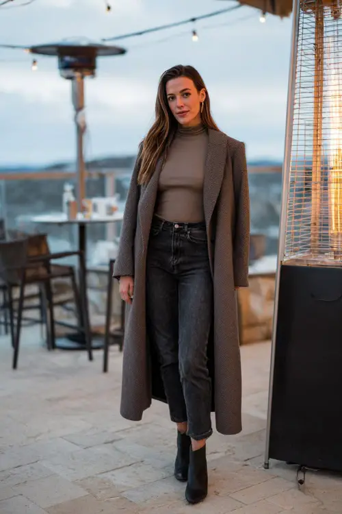 A woman wears a long-sleeve fitted top under a wool wrap coat, paired with dark denim and block-heel ankle boots, standing near an outdoor patio heater on a winter dinner date