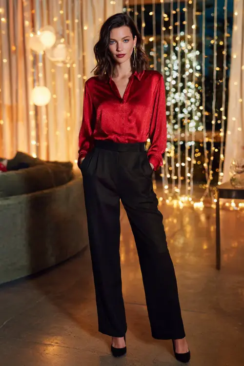 A woman wears a red silk blouse with subtle shimmer tucked into wide black trousers, delicate earrings, and black heels, standing full body in a festive living room party with soft string lights 