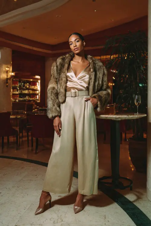 A woman wears a silky champagne blouse tucked into high-waisted wide-leg pants with a belted waist, paired with heeled pumps and a cropped faux-fur jacket