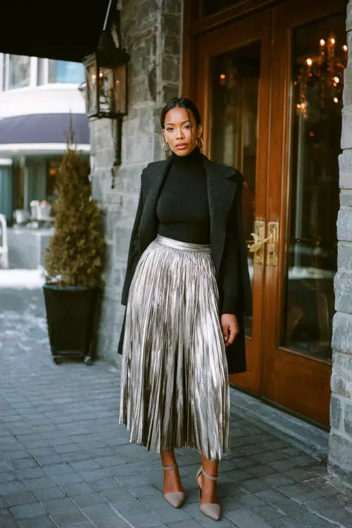 A woman wears a sleek black mock-neck top tucked into a shimmering metallic pleated skirt, styled with ankle-strap heels and a wool wrap coat