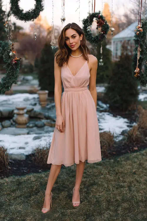 A woman wears a soft pastel pink chiffon midi dress, minimalist pearl necklace, satin heels, posed full body in a dreamy winter garden decorated with hanging Christmas wreaths