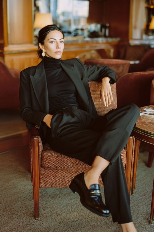 A woman wears a tailored black blazer over a fitted turtleneck with high-waisted black trousers and polished leather loafers