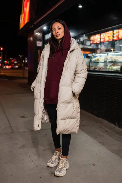 A woman wears a warm burgundy hoodie under a long puffer coat, styled with leggings and chunky winter sneakers, standing near a casual nighttime food spot