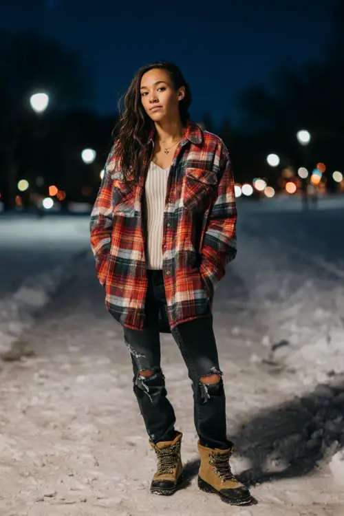 A woman wears an oversized plaid shacket over a ribbed top, paired with distressed jeans and lace-up winter boots, posing on a snowy park path at dusk